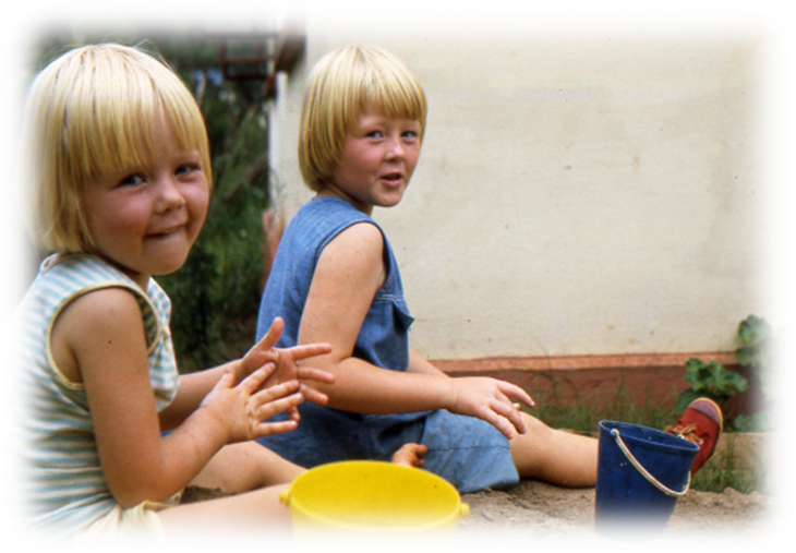 Two young girls playing in sand; one wears a striped shirt and smiles at the camera, while the other wears a blue dress and looks back. They have short blonde hair and bright yellow and blue buckets beside them.